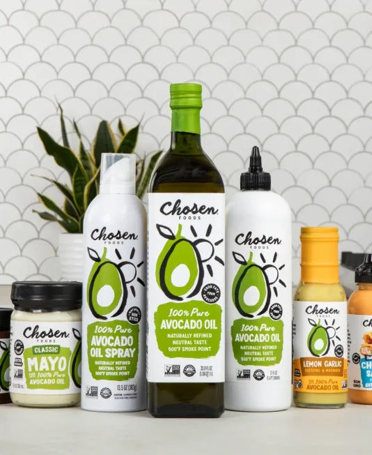 A lineup of Chosen Foods products against a tiled wall: avocado oil bottles and spray, classic mayonnaise, and lemon garlic dressing.