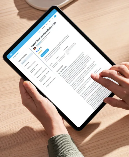 Two hands holding a tablet on a wooden desk, displaying a document titled Supply Chain Operations Buying Guide.